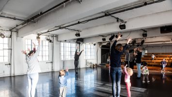 In a large dance studio, a group of parents and their children reach their hands up and lean to one side.