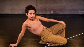 Mariana reclines on a dark stage, with her hand on her knee