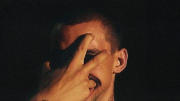James Batchelor's fingers press into his face, on a dark background