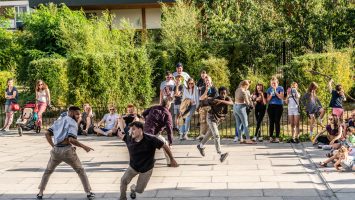 Four dancers from different ethnic backgrounds running and smiling in the centre of a large square. The audience are spread along the edges watching and smiling.