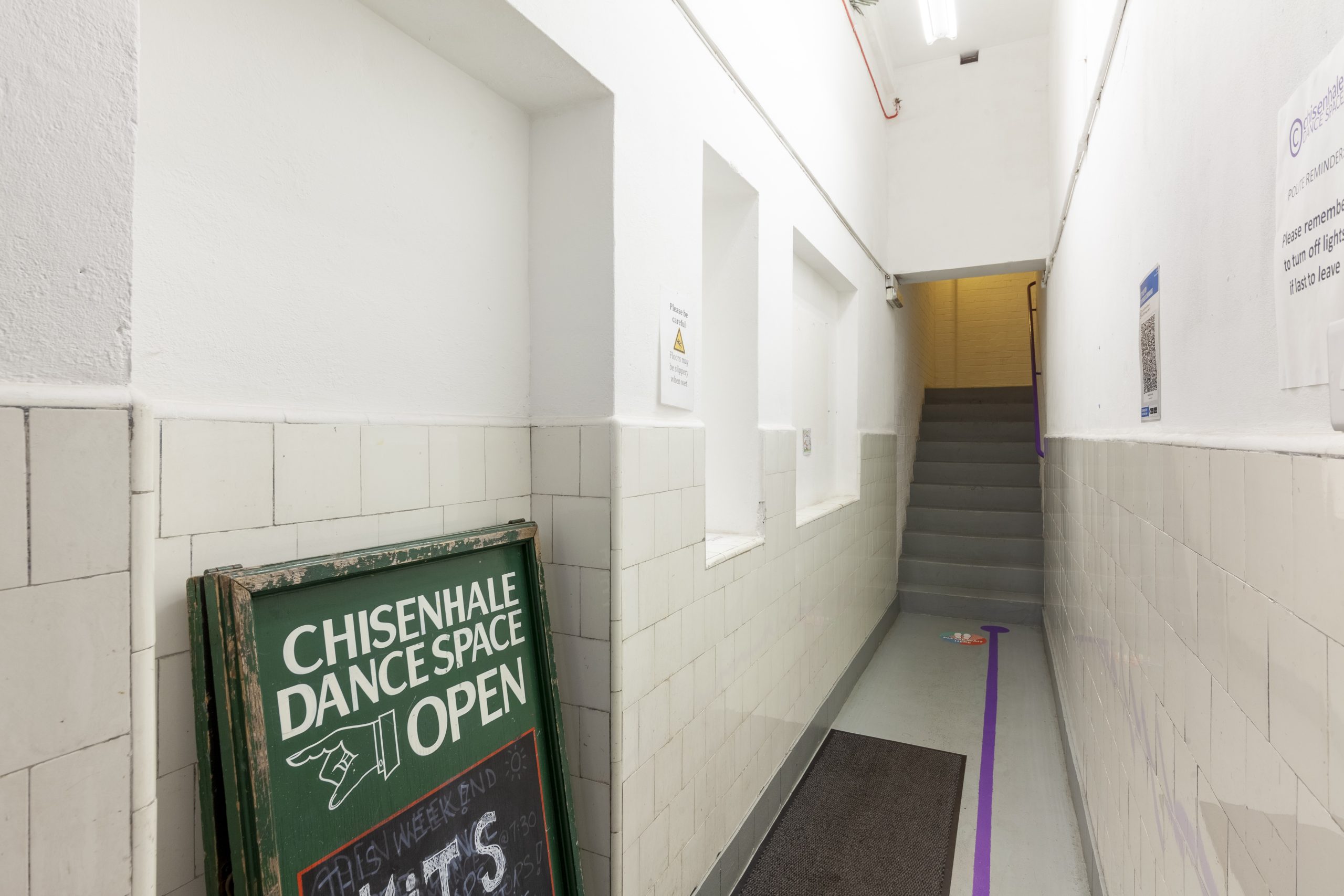 The entrance to a building painted white, with a purplue line on the floor leading to some stairs going upwards