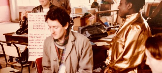 a sepia image of four smiling people gathered in a warehouse space, preparing for a protest. They are playing a keyboard and holding a banner with chants on it