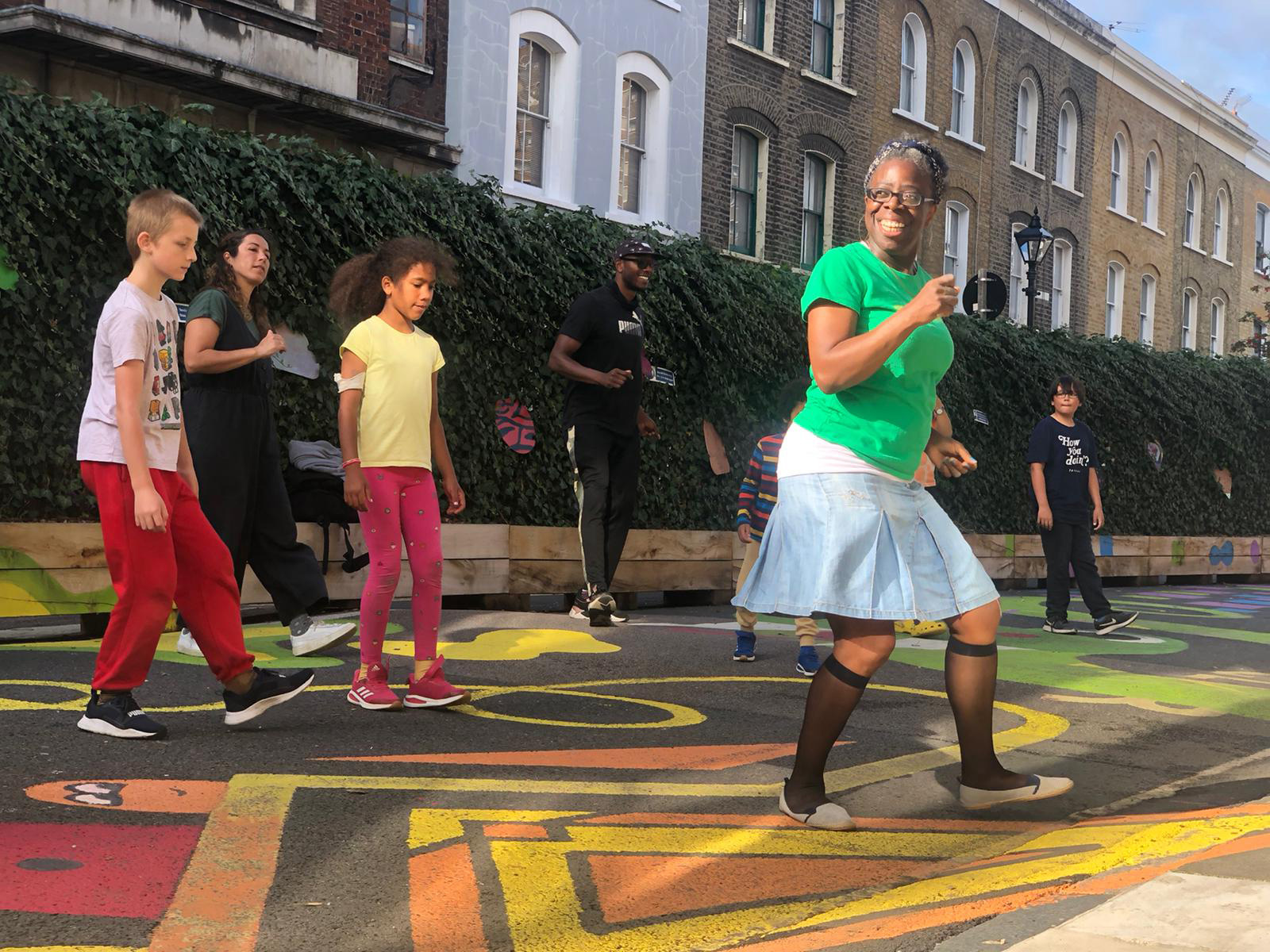 A black woman wearing a bright green t-shirt and a white skirt is leading an outdoor Lindy Hop class for children and adults.