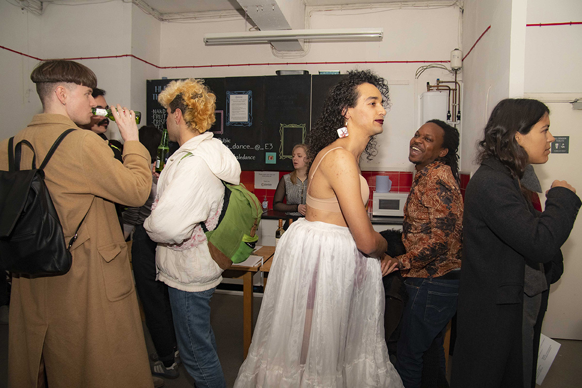 A group of people are gathered in the Chisenhale Dance Space communal area. A medium skinned performer with long curly hair is moving out of frame. They are wearing white dangly earrings, a bra that matches their skin tone and a white lace skirt. A dark skinned man in a patterned shirt looks on smiling widely.