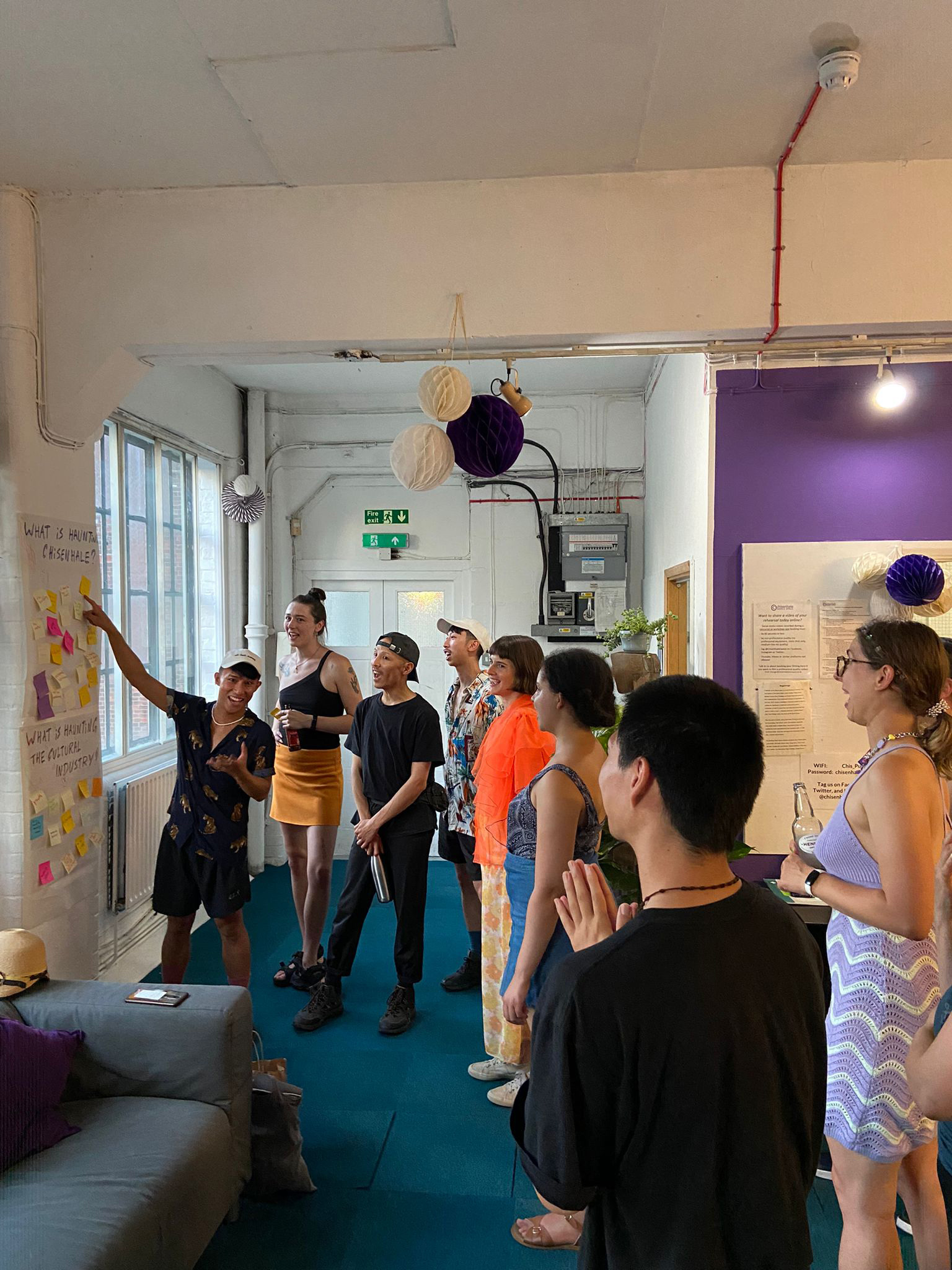 An East-Asian man is pointing towards a large piece of paper with various coloured sticky notes attached. He is looking towards a group of participants. They are in the communal area of Chisenhale Dance Space.