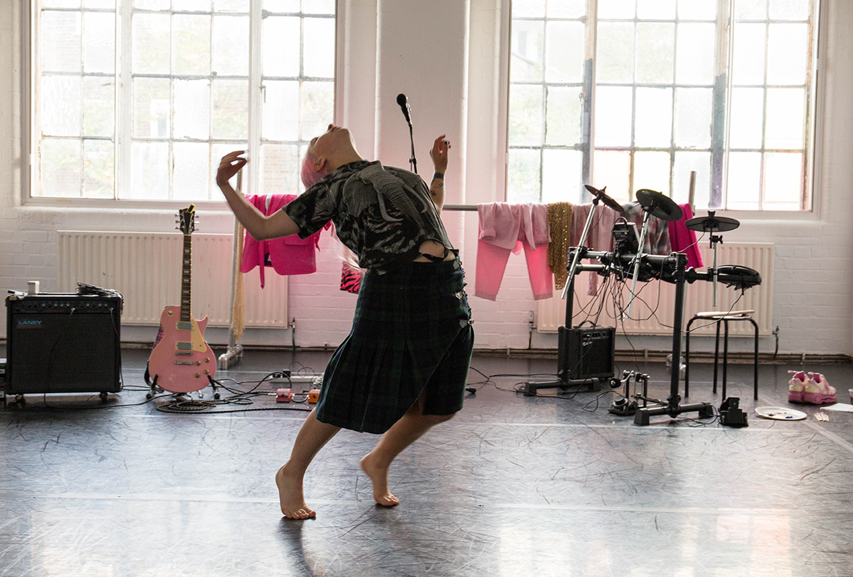 A light skinned person with pink hair is pictured arching their back while off-balance in a dance studio. There are various pink items of clothing hung on a ballet barre in the background as well as a pink electric guitar and drum kit.