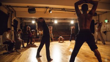 An image of a movement workshop where attendees are stood in a circle, some stretching upwards, some crouched down. There are two musicians playing drums to the side of the space.