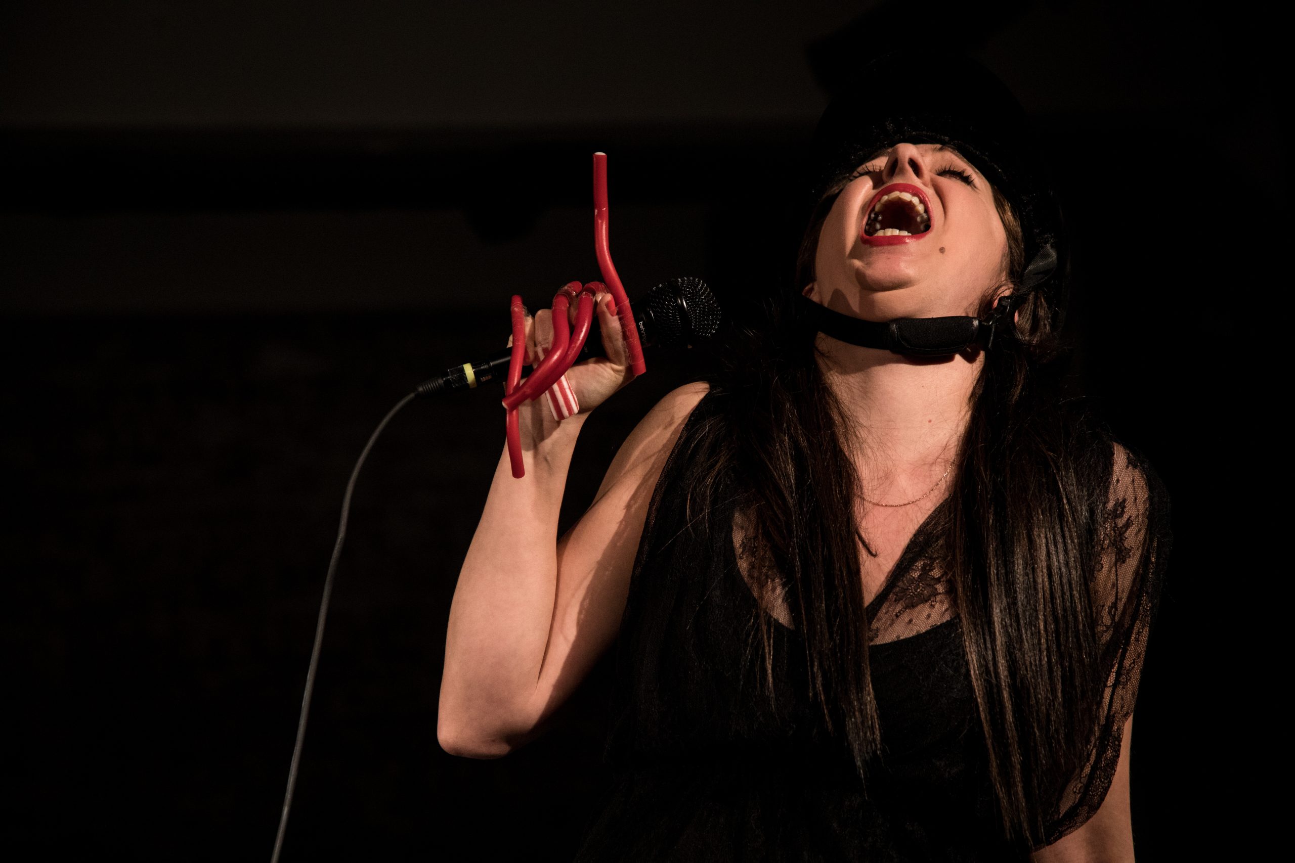 A white woman is holding a microphone, head tilted back. She has strawberry pencils taped to each finger in her right hand and a black strap under her chin.