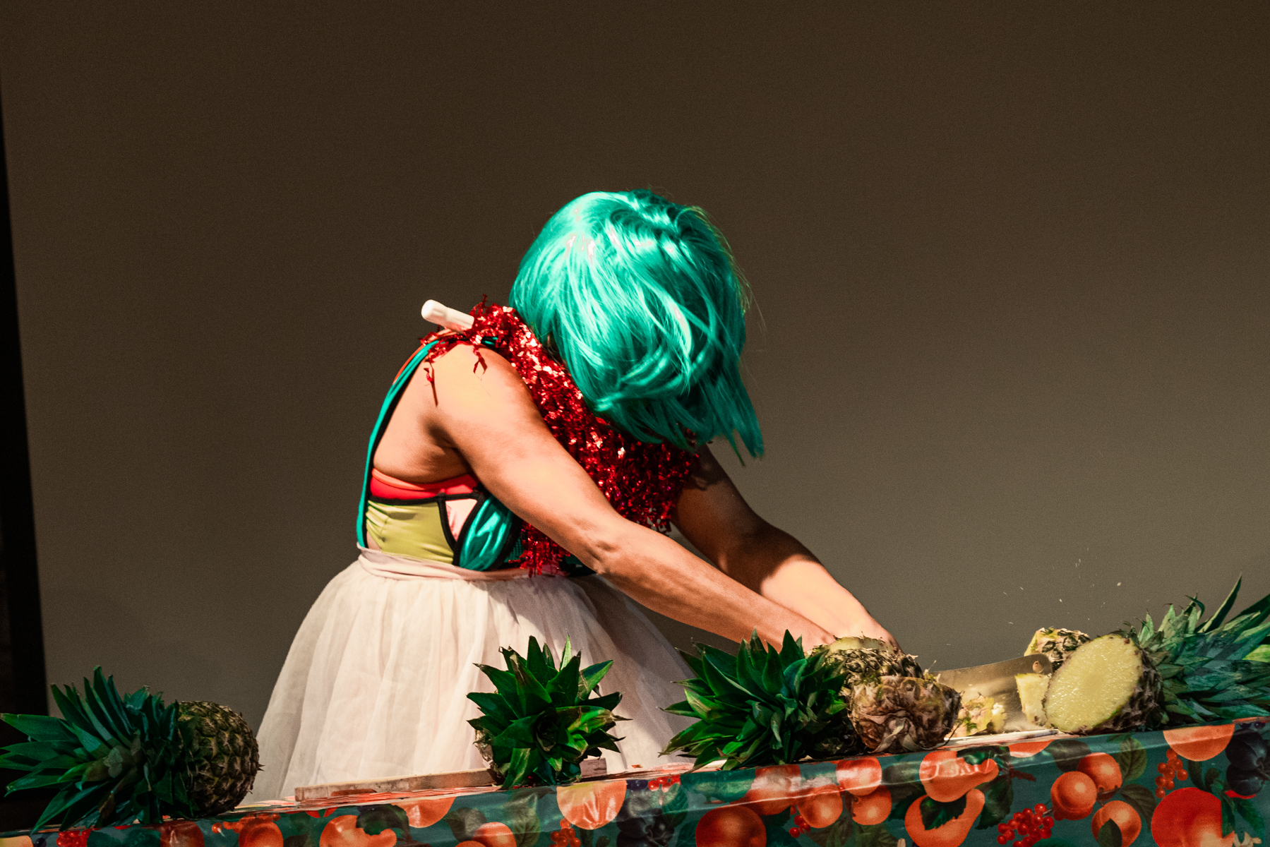 A medium-skinned performer in a turquoise wig and a white tutu cuts a pineapple in half with a cleaver onstage. There are several other pineapples on the table in various states of wholeness.